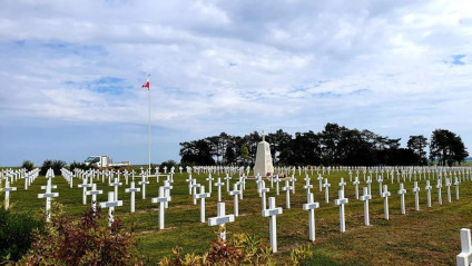 Polski Cmentarz Wojenny w Auberive z rzędami białych krzyży i centralnym pomnikiem. Na wysokim maszcie powiewa polska flaga. W tle drzewa pod pochmurnym niebem.