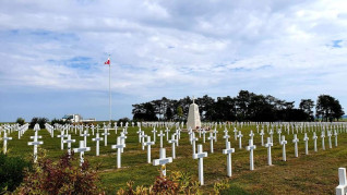Polski Cmentarz Wojenny w Auberive z rzędami białych krzyży i centralnym pomnikiem. Na wysokim maszcie powiewa polska flaga. W tle drzewa pod pochmurnym niebem.