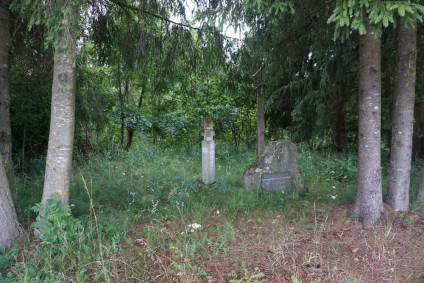 Cmentarz powstańców styczniowych otoczony drzewami. Widoczne są słup kapliczny, betonowy krzyż i głaz z litewską inskrypcją.