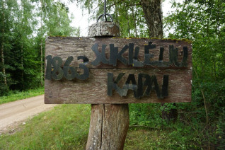 Drewniany znak z napisem '1863 Suklėlių Kapai' w leśnym otoczeniu, wskazujący lokalizację cmentarza powstańców styczniowych.