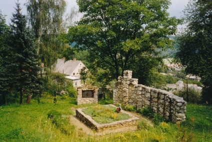 Kamienny pomnik z krzyżem i symbolicznymi postaciami na trawiastym miejscu pochówku, otoczony drzewami i domem w tle, upamiętniający Polaków zamordowanych przez Niemców pod Krzemieńcem.