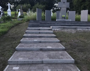 Kamienne schody prowadzą do miejsca pamięci z centralnym krzyżem i pionowymi tablicami otoczonymi zielenią. Miejsce upamiętnia polskie ofiary masakry z 1944 roku w Palikrowach.