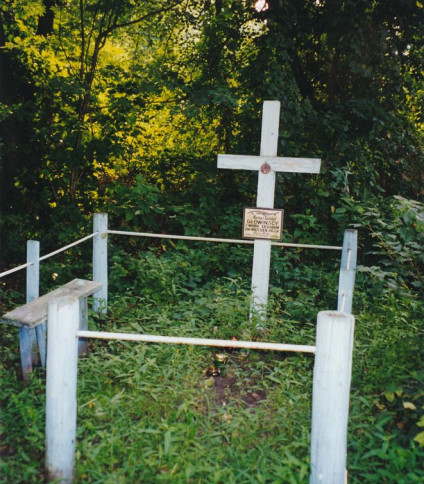 Biały krzyż z tabliczką stoi na trawiastym terenie otoczonym prostym drewnianym ogrodzeniem, oznaczając grób polskich ofiar UPA w zalesionej okolicy.