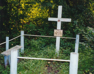 Biały krzyż z tabliczką stoi na trawiastym terenie otoczonym prostym drewnianym ogrodzeniem, oznaczając grób polskich ofiar UPA w zalesionej okolicy.