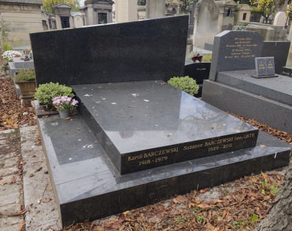 Nagrobek Karola i Suzanne Barczewskich z pochyloną, schodkową podstawą i prostokątną stelą z krzyżem po prawej. Widoczne są inskrypcje z przodu i z boku. Na grobie umieszczono kwiaty.