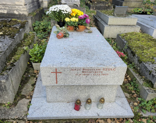 Grób Jerzego Szulca z dużą kamienną płytą na wysokim cokole. Na froncie inskrypcja i krzyż. Na wierzchu kwiaty i znicze.