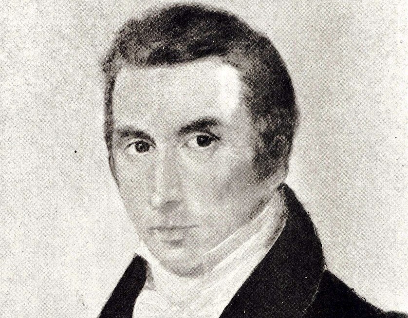 Portrait d'un homme aux cheveux noirs, portant un manteau sombre et un nœud papillon blanc, regardant droit dans les yeux.