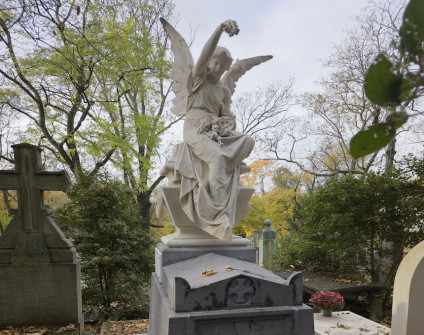 Marmurowy nagrobek z siedzącym aniołem trzymającym kwiaty. Na froncie nagrobka krzyż wpisany w koło. Scenę otaczają drzewa z jesiennymi liśćmi.