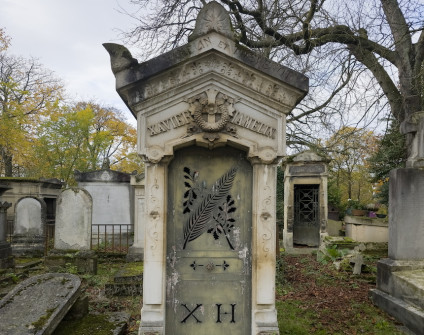 Kapliczka klasycystyczna z dwuspadowym dachem, akroteriony na rogach i szczycie, fryz z motywem roślinnym, krzyż z wieńcem i szarfami w tympanonie, drzwi metalowe z wzorem gałęzi dębu i palmy, na dole wycięte litery 'X H'.