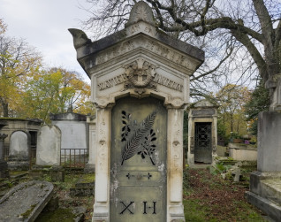 Kapliczka klasycystyczna z dwuspadowym dachem, akroteriony na rogach i szczycie, fryz z motywem roślinnym, krzyż z wieńcem i szarfami w tympanonie, drzwi metalowe z wzorem gałęzi dębu i palmy, na dole wycięte litery 'X H'.