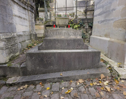 Nagrobek rodziny Wagner na schodkowym cokole z wyrytą inskrypcją 'FAMILLE WAGNER'. U wezgłowia kamienna żardiniera z czerwonymi świecami. Opadłe liście pokrywają brukowaną ziemię.