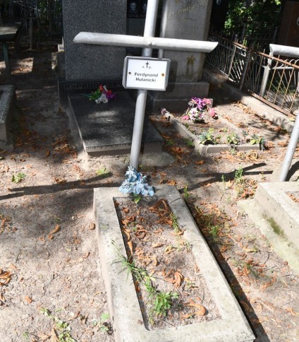 Grób oznaczony rurkowym krzyżem stalowym z metalową tabliczką z napisem 'Ferdynand Hulanicki'. Grób otoczony kamienną opaską z roślinami i opadłymi liśćmi.