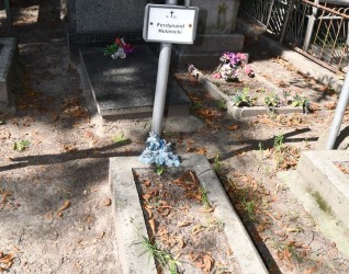 Grób oznaczony rurkowym krzyżem stalowym z metalową tabliczką z napisem 'Ferdynand Hulanicki'. Grób otoczony kamienną opaską z roślinami i opadłymi liśćmi.