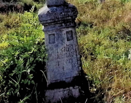 Nagrobek Bronisława i Stanisława Skurskich z krzyżem na dwustopniowym cokole. Krzyż z postacią Chrystusa i inskrypcją poniżej. Otoczony trawą i roślinami.