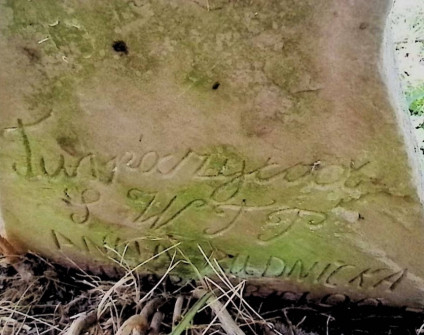 Zbliżenie na nagrobek z wyrytą inskrypcją częściowo zasłoniętą trawą. Kamień ma zniszczoną powierzchnię.