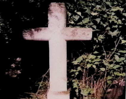 Kamienny krzyż o fazowanych ramionach na cokole, otoczony gęstą zielenią.