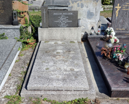 Nagrobek rodziny Krucinów z inskrypcją na steli na cokole. Z lewej przylega prostopadłościenny bloczek z rytym krzyżem. Grób otoczony innymi nagrobkami i zielenią.