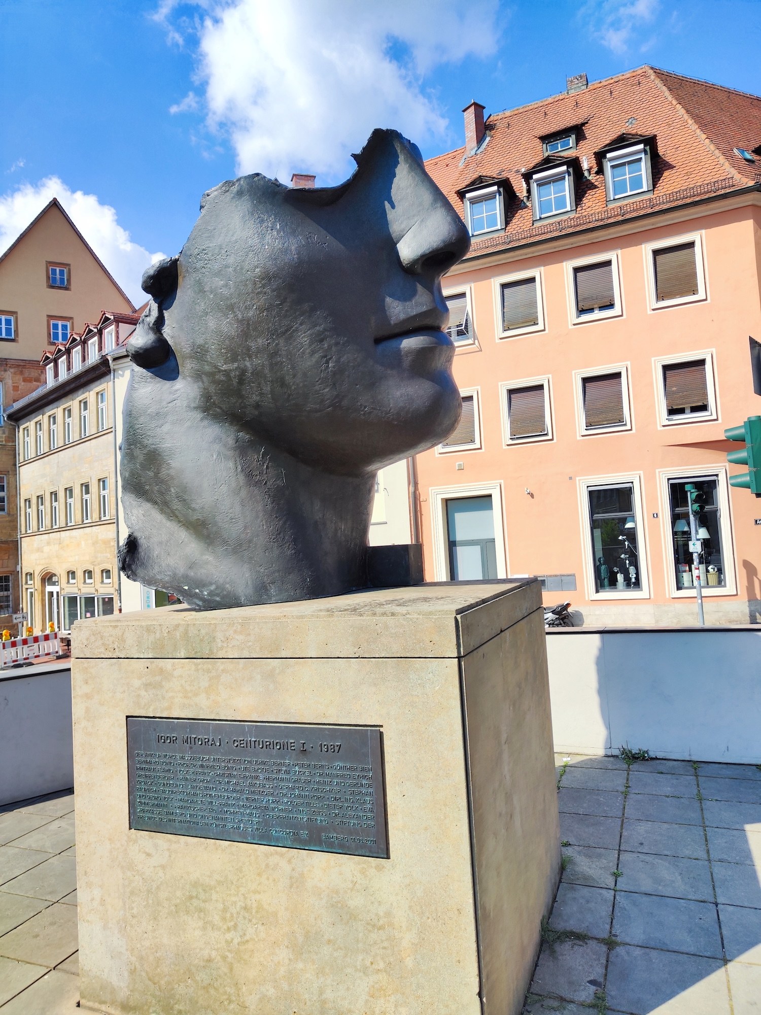 Bronze sculpture 'Centurione' by Igor Mitoraj in Bamberg, depicting a fragmented male head with a classic nose and lips, set on a pedestal with an inscription plaque. Background shows traditional European buildings.