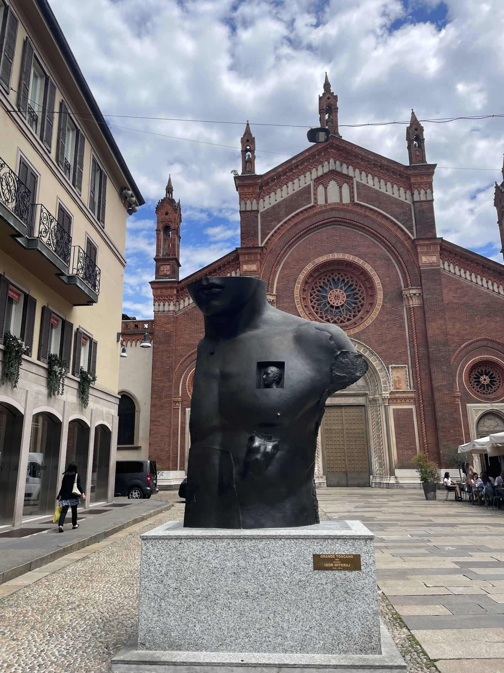 Bronze sculpture 'Grande Toscano' by Igor Mitoraj in Milan, depicting a fragmented male torso with a window containing a male head and a female torso. Located in front of a red brick church.