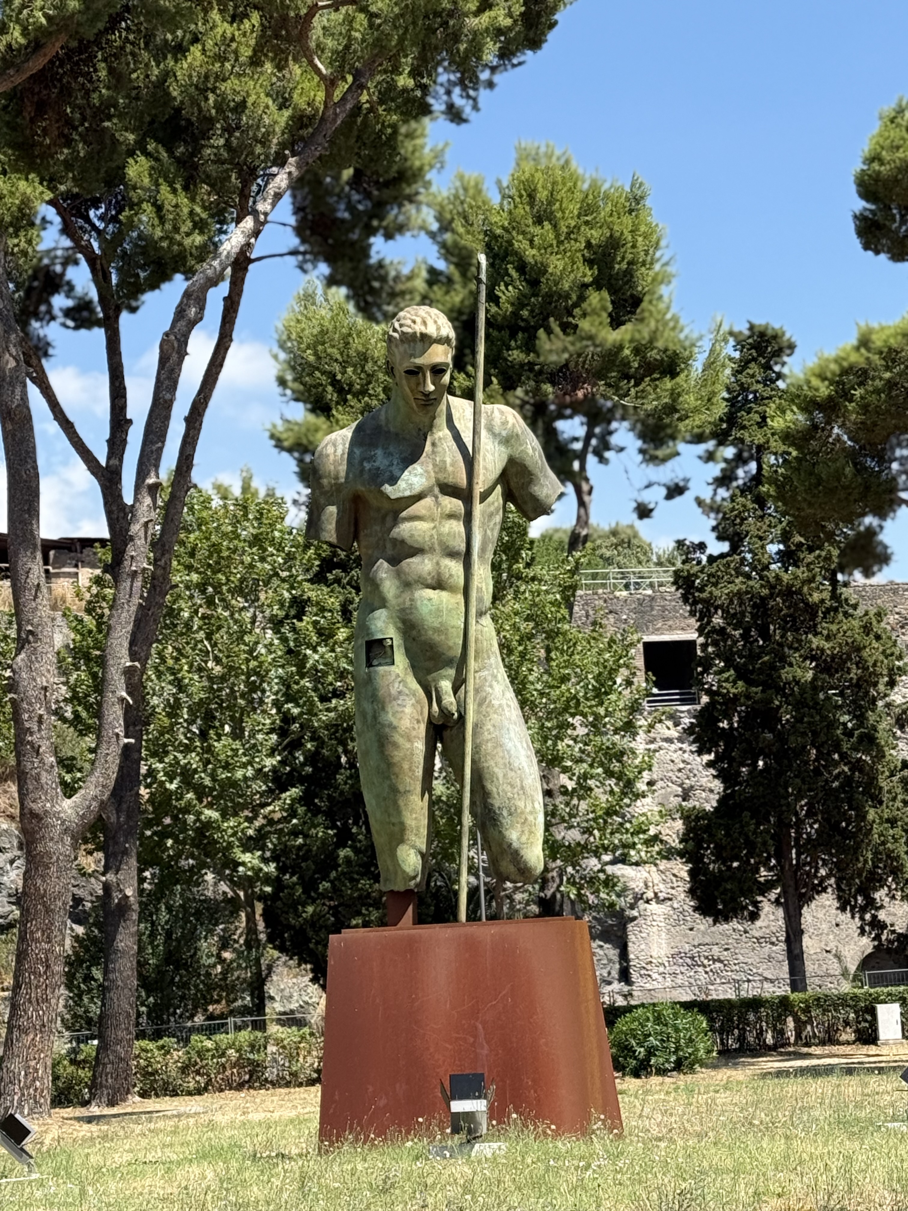Bronze sculpture of a male torso by Igor Mitoraj, missing arms, standing on a rust-colored pedestal. The figure holds a long spear. Trees and a stone wall are in the background.