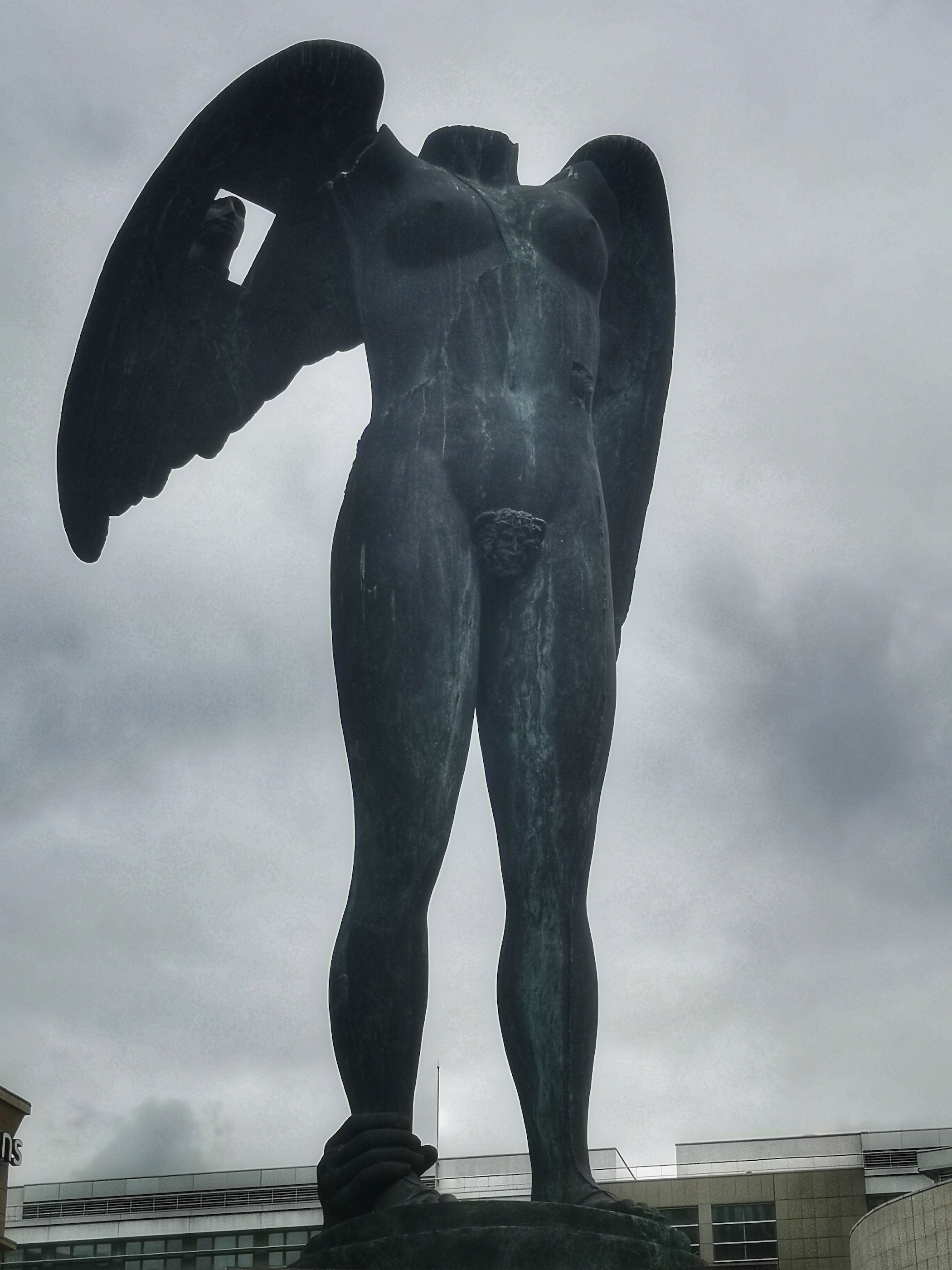 Bronze sculpture 'Ikaria' by Igor Mitoraj in Paris. It depicts a headless female figure with wings, standing on a pedestal. The right foot is held by a hand, and a small female figure is attached to the left ankle.