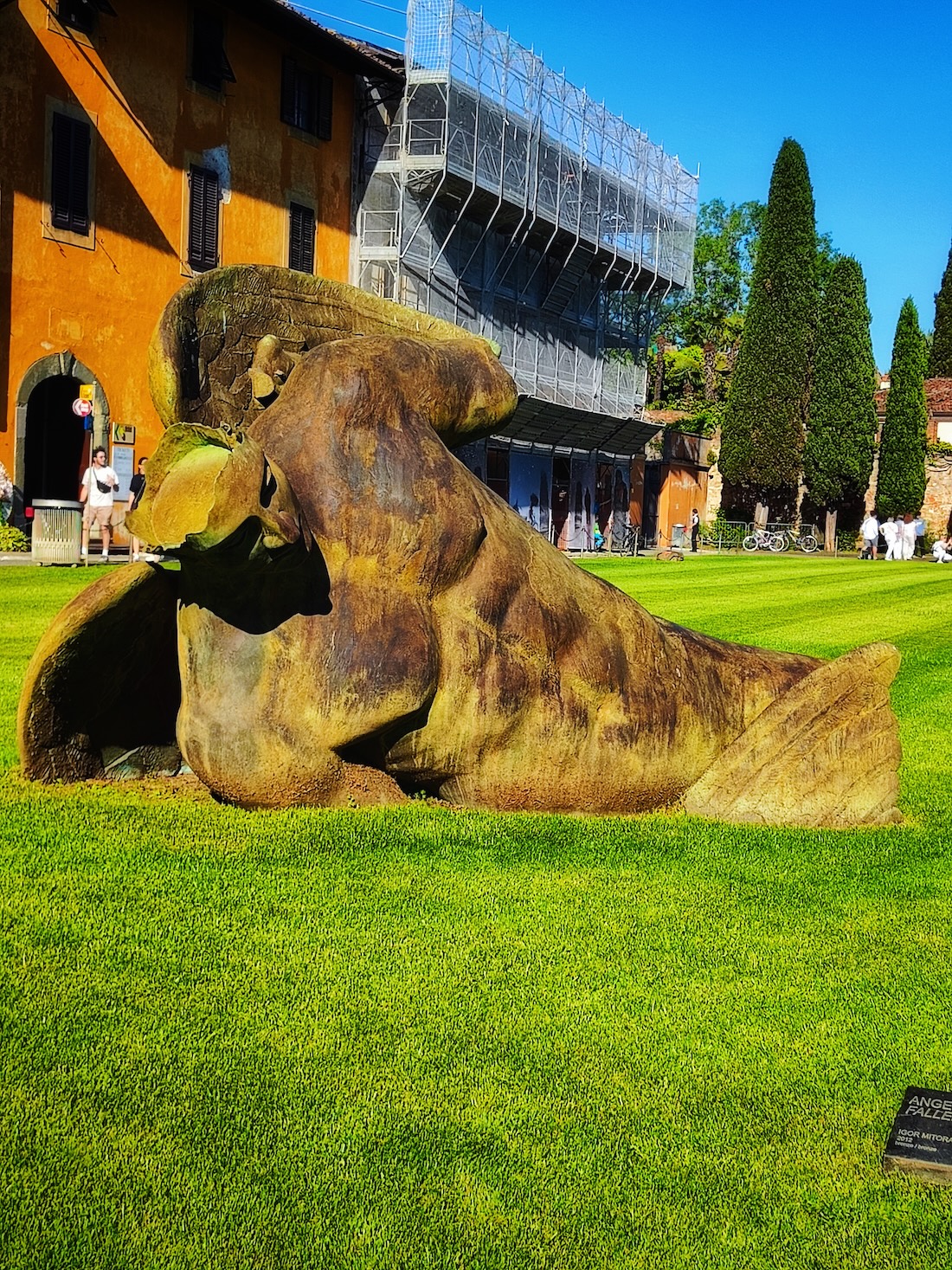 Bronze sculpture 'Fallen Angel' by Igor Mitoraj on the grass at Piazza dei Miracoli in Pisa, Italy. The sculpture depicts a winged male torso without a pedestal. In the background, an orange building and trees.