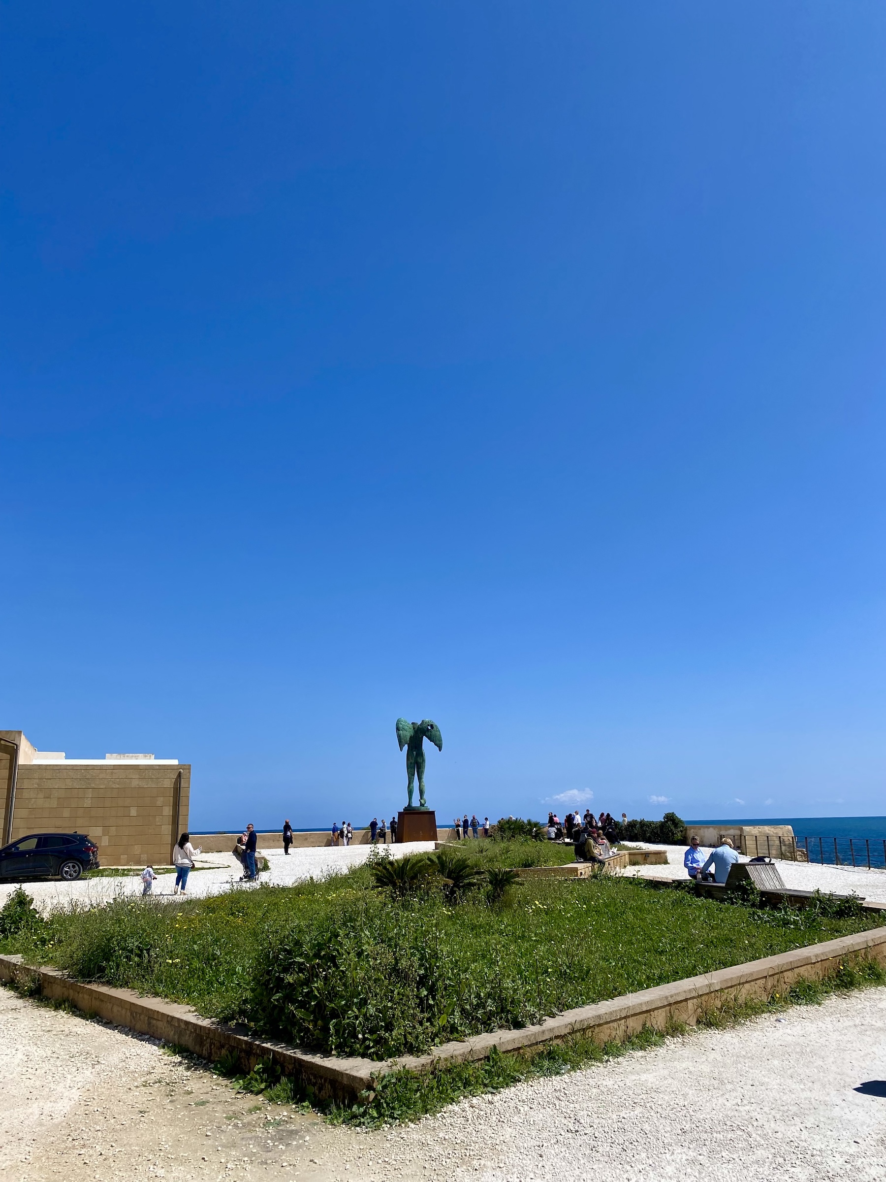 The sculpture 'Upadek Ikara' by Igor Mitoraj in Agrigento, Italy, depicts a winged male torso on a pedestal. Visitors walk around the sculpture, set against a clear blue sky and sea.