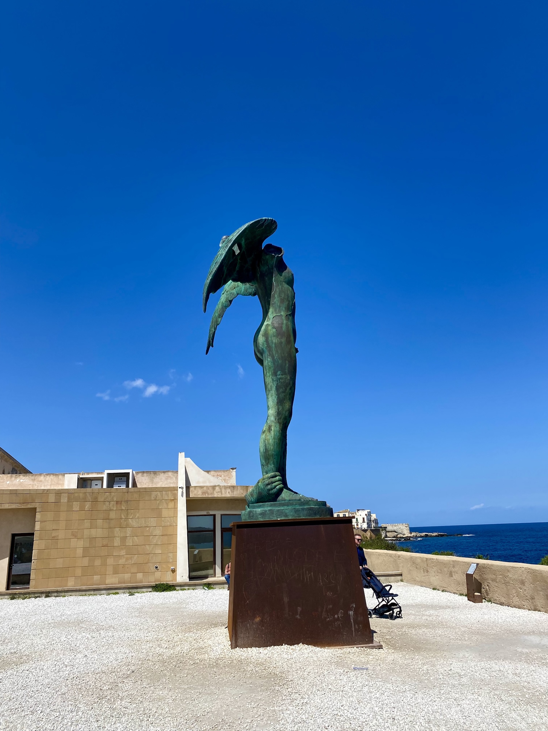 Bronze sculpture 'Icarus' by Igor Mitoraj in Syracuse, Sicily. The headless, winged figure stands on a pedestal against a blue sky and sea backdrop.