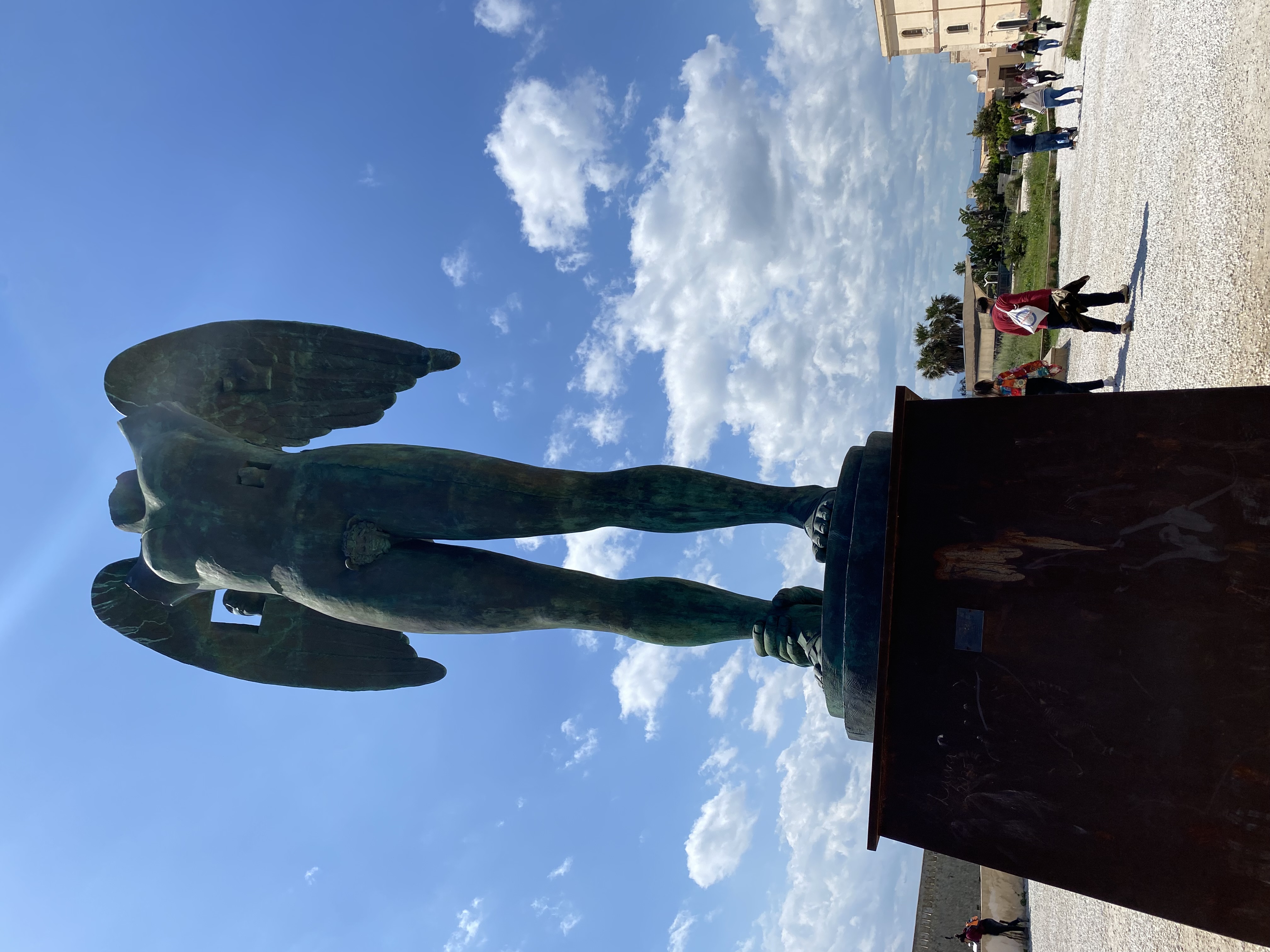 Bronze sculpture of a headless, winged male torso on a pedestal, set against a blue sky with scattered clouds. People walk in the background on a gravel path.