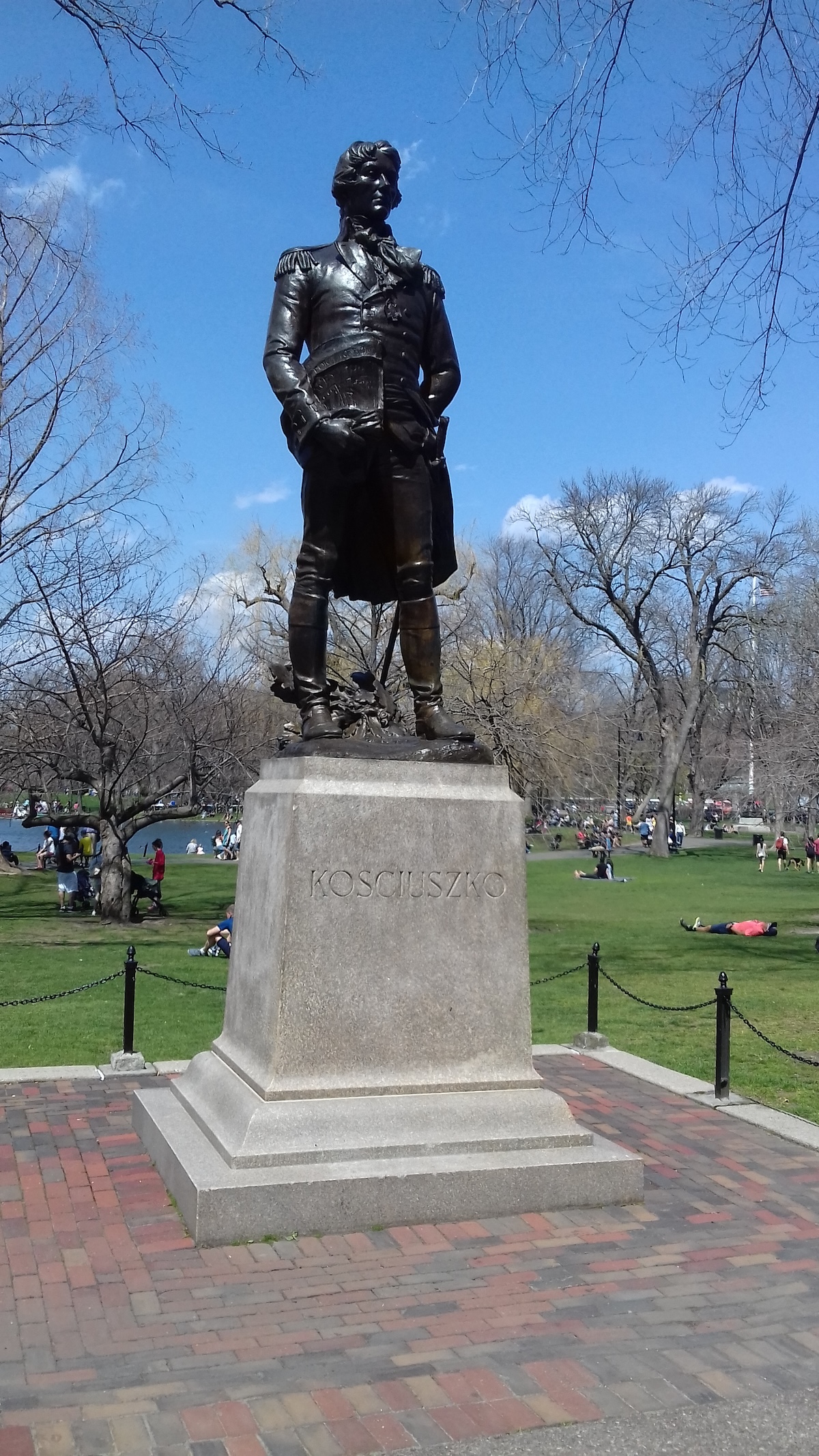 Statue en bronze de Tadeusz Kościuszko dans le Public Garden de Boston sur un socle en granit, entourée d'arbres et de personnes par une journée ensoleillée.