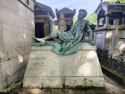 Brązowa rzeźba leżącego mężczyzny w mundurze na cokole w formie sarkofagu, z inskrypcjami i dekoracjami. Znajduje się na cmentarzu otoczonym innymi grobami.