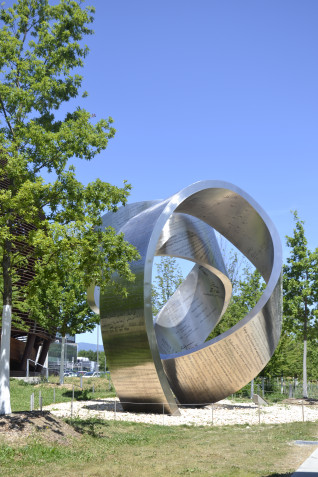 Stainless steel sculpture 'Wandering the Immeasurable' at CERN, featuring a twisting ribbon with inscriptions of scientific discoveries, including names of Marie Curie and Hermann Minkowski. Surrounded by trees and grass.