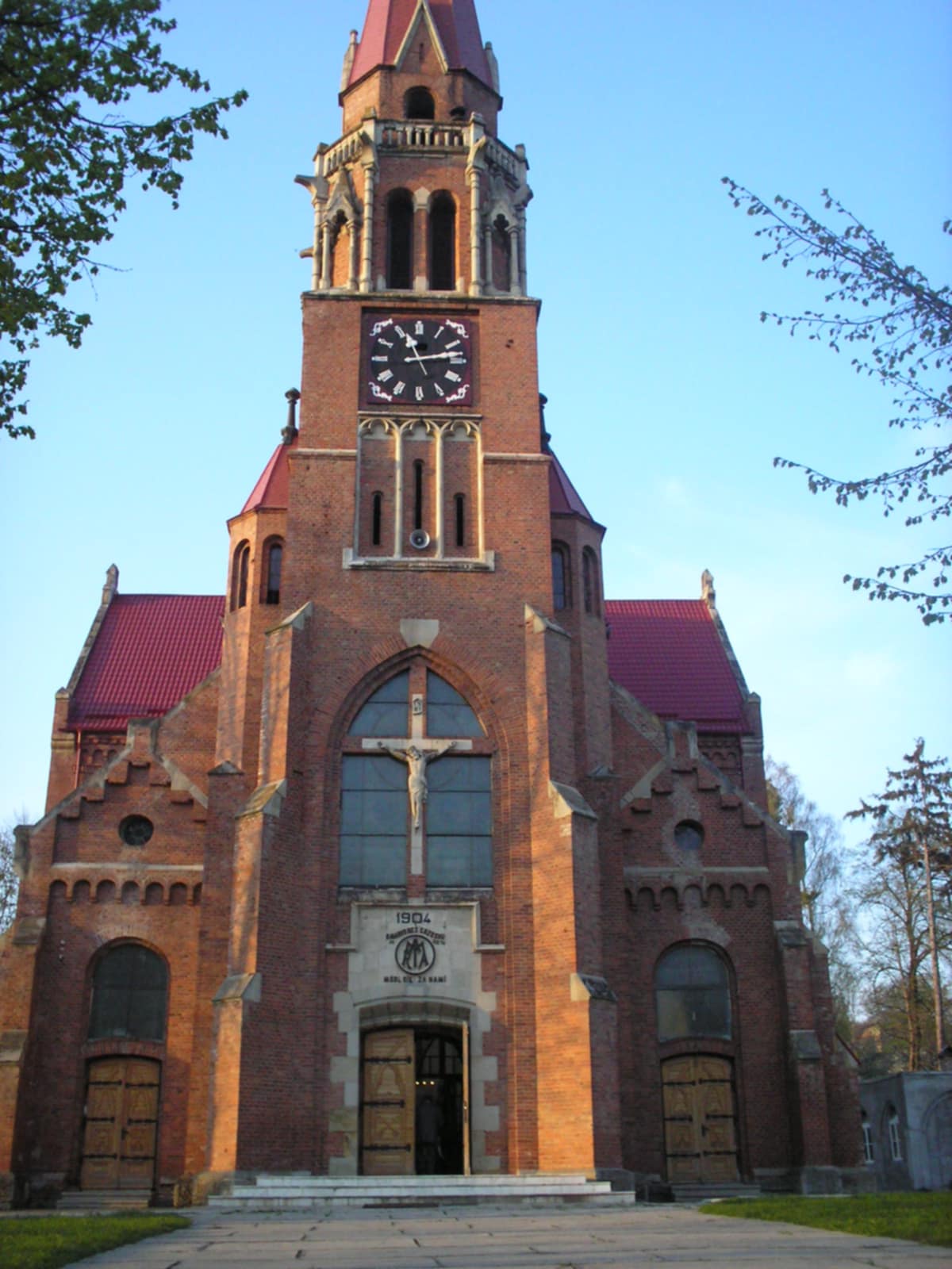 The Basilica of the Assumption of the Blessed Virgin Mary in Cacica, Romania, built in 1904. The brick facade features a large clock tower, arched windows, and a crucifix above the entrance.