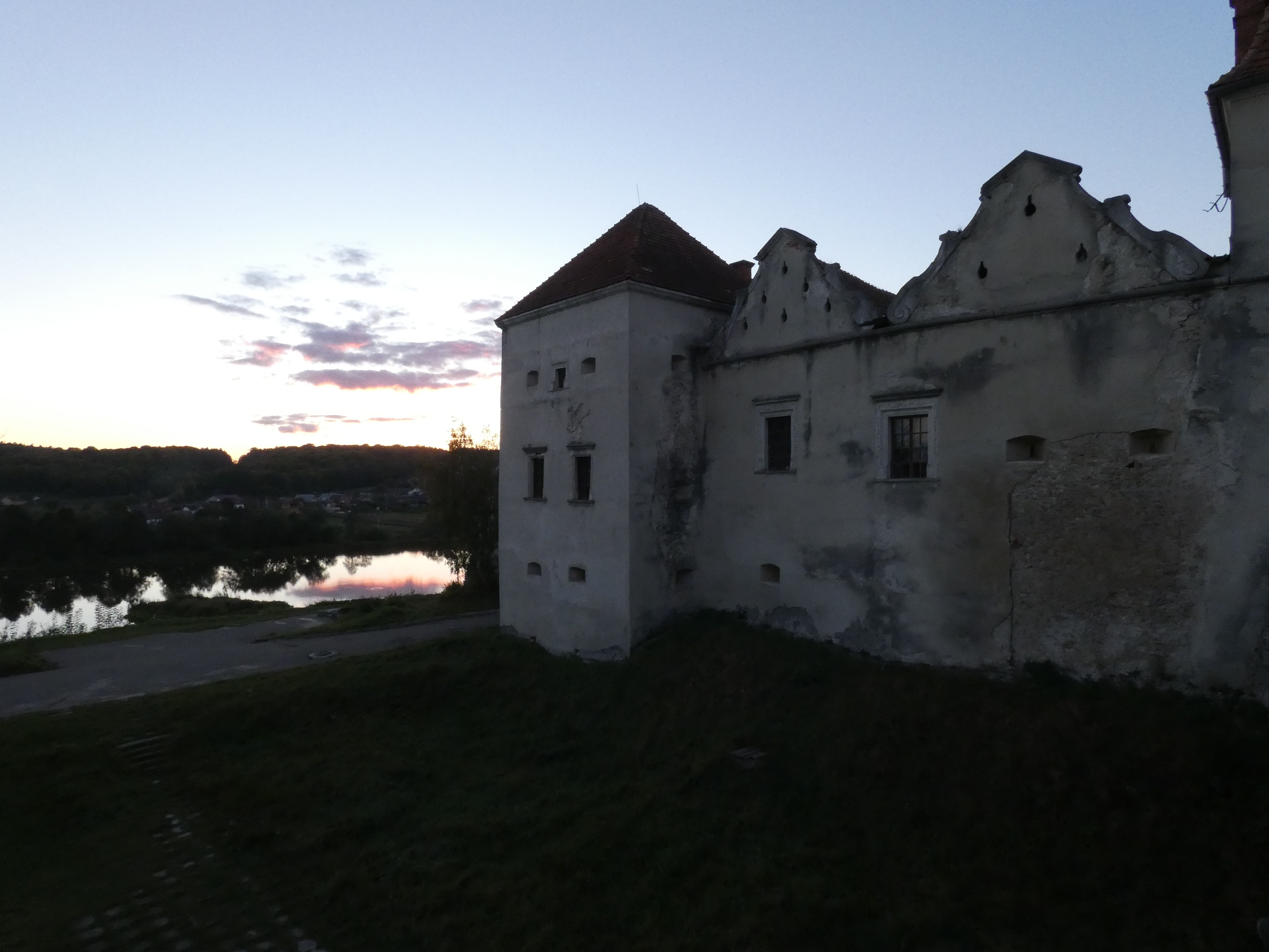 Swirz castle at dusk overlooking the lake and surrounding landscape. The building has decorative gables and small windows, with signs of wear on the walls.