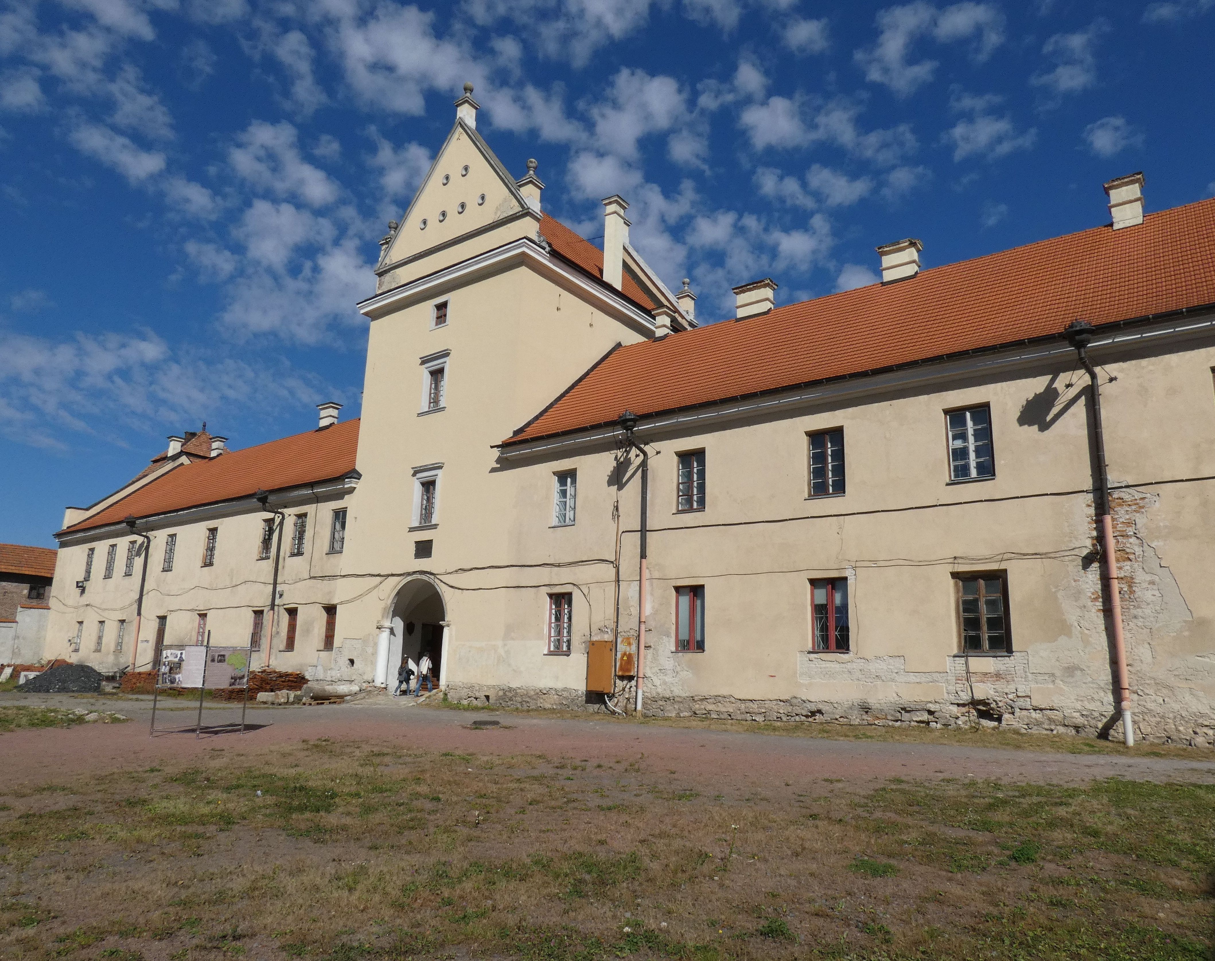 Фасад замку в Жовкві, Україна, з бежевою будівлею з трикутним фронтоном і червоним дахом. Будівля має багато вікон та арочний вхід. Небо ясне з розсіяними хмарами.