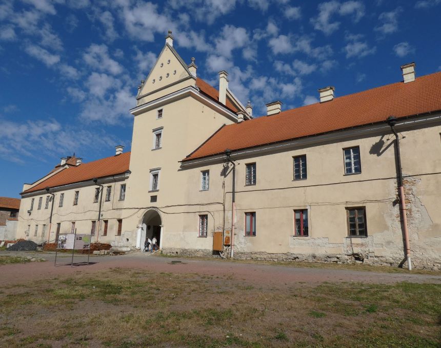 Фасад Жовківського замку з бежевими стінами та червоним дахом на тлі синього неба з хмарами. Будівля має багато вікон і центральну вхідну арку.