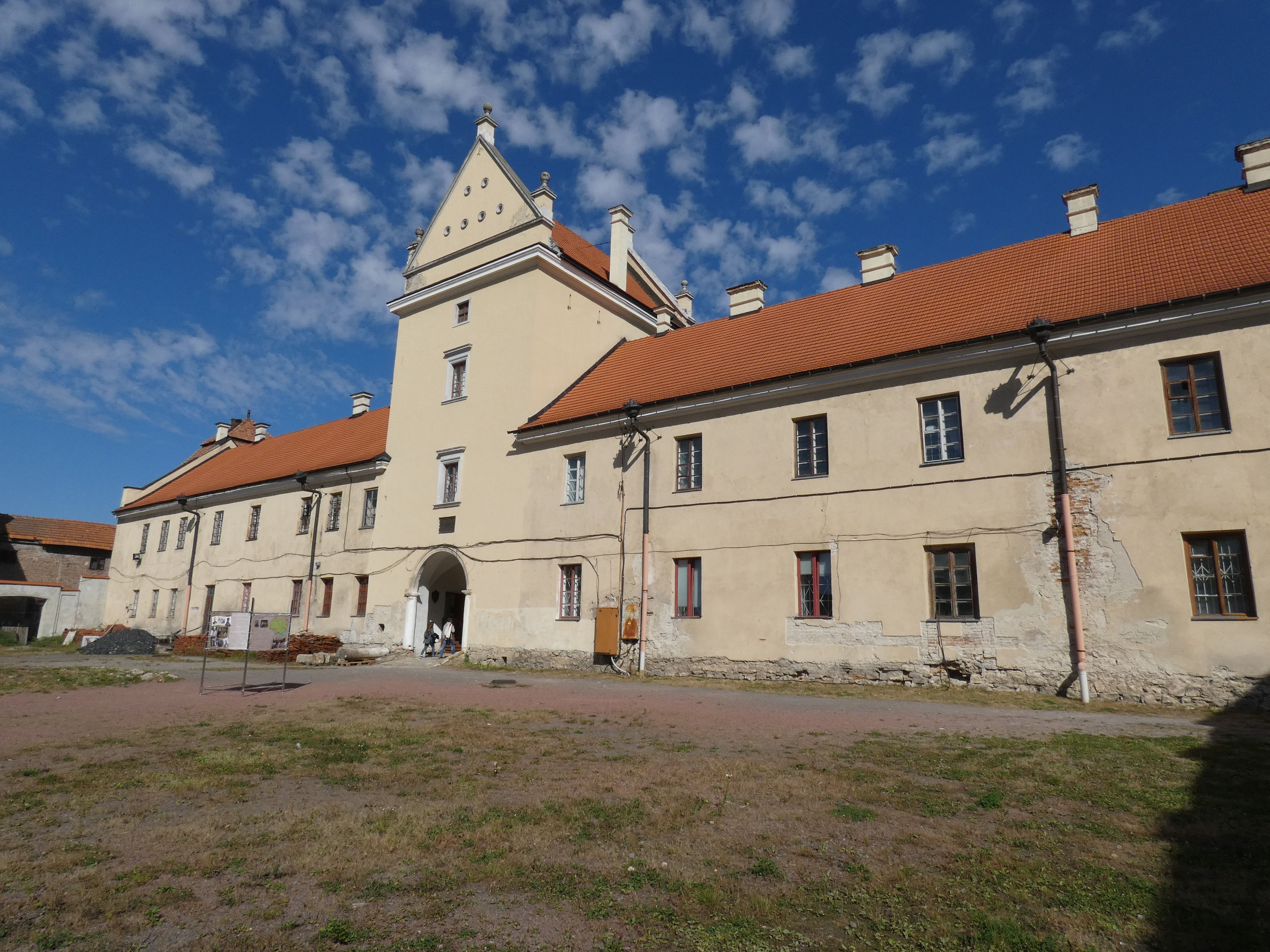 Фасад замку в Жовкві з бежевими стінами та червоним дахом під блакитним небом з хмарами. Будівля має багато вікон і центральну арку входу.