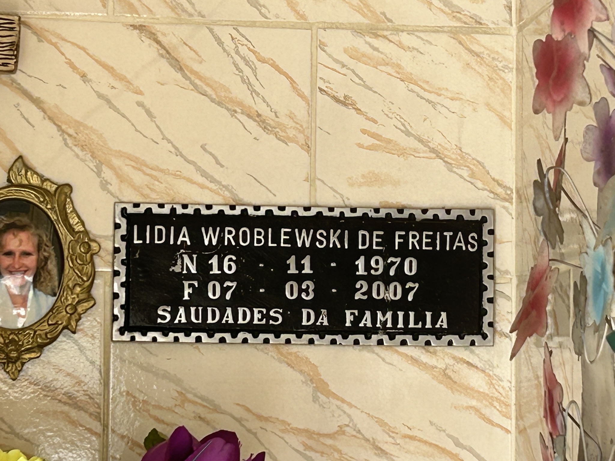 Plaque portant l'inscription "Lidia Wroblewski de Freitas, N 16-11-1970, F 07-03-2007, Saudades da Familia" sur un mur carrelé. À gauche, une photographie encadrée d'une femme.