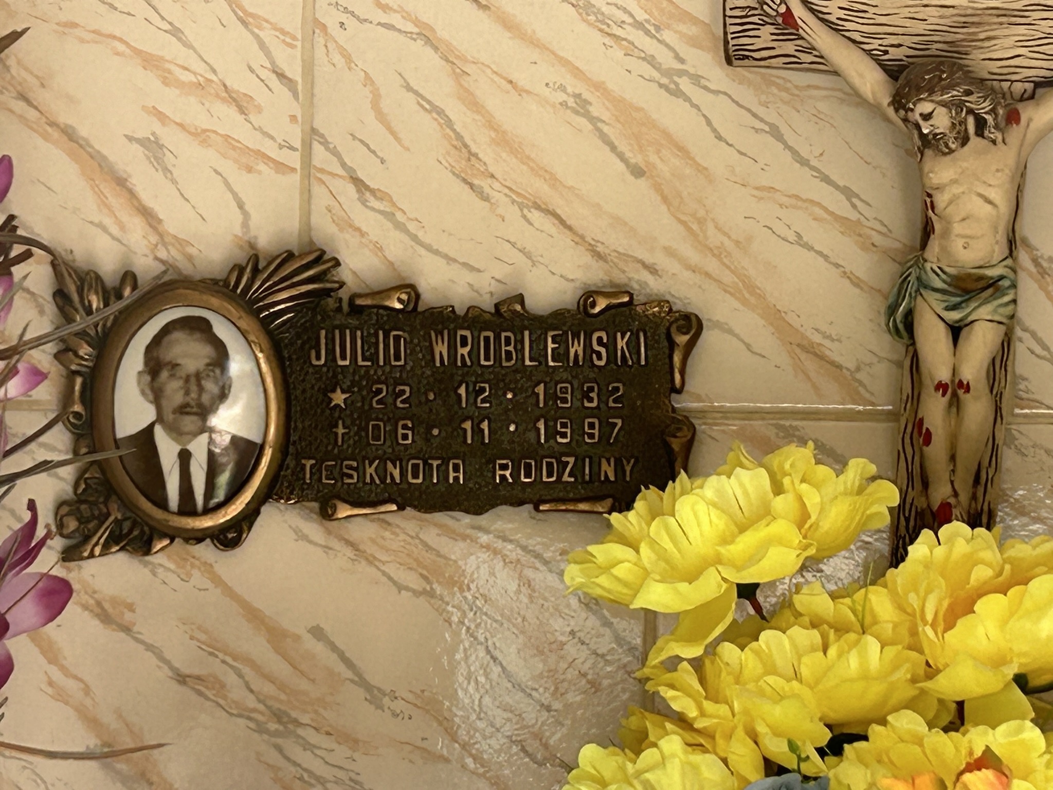 Plaque avec une photographie de Juliusz Wróblewski, les dates 22.12.1932 et 06.11.1997 et l'inscription "Longing for the Family". À côté d'un crucifix et de fleurs jaunes.