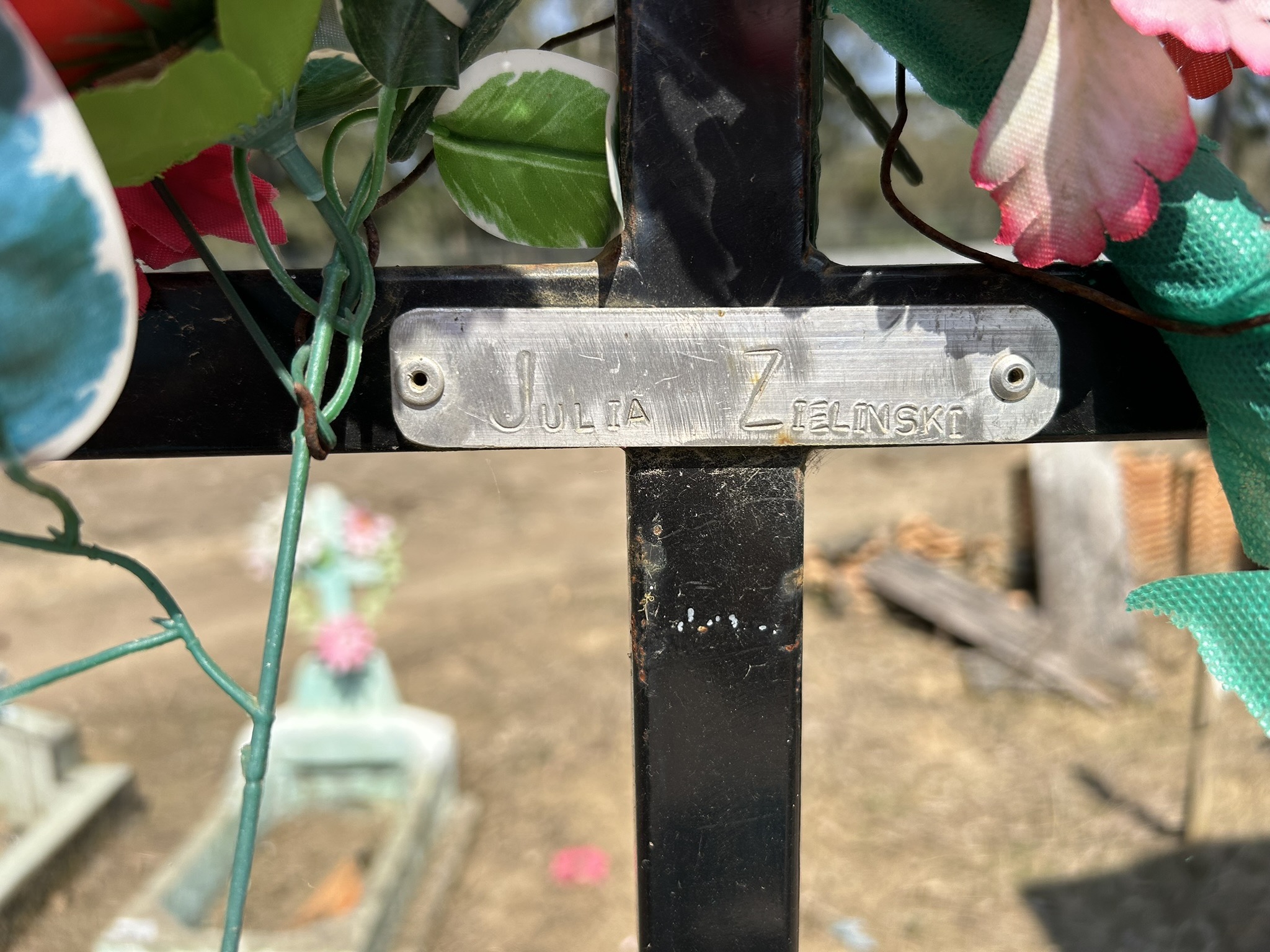 Gros plan d'une plaque métallique sur une croix funéraire portant l'inscription "Julia Zielinski". Fleurs artificielles de différentes couleurs autour de la plaque.
