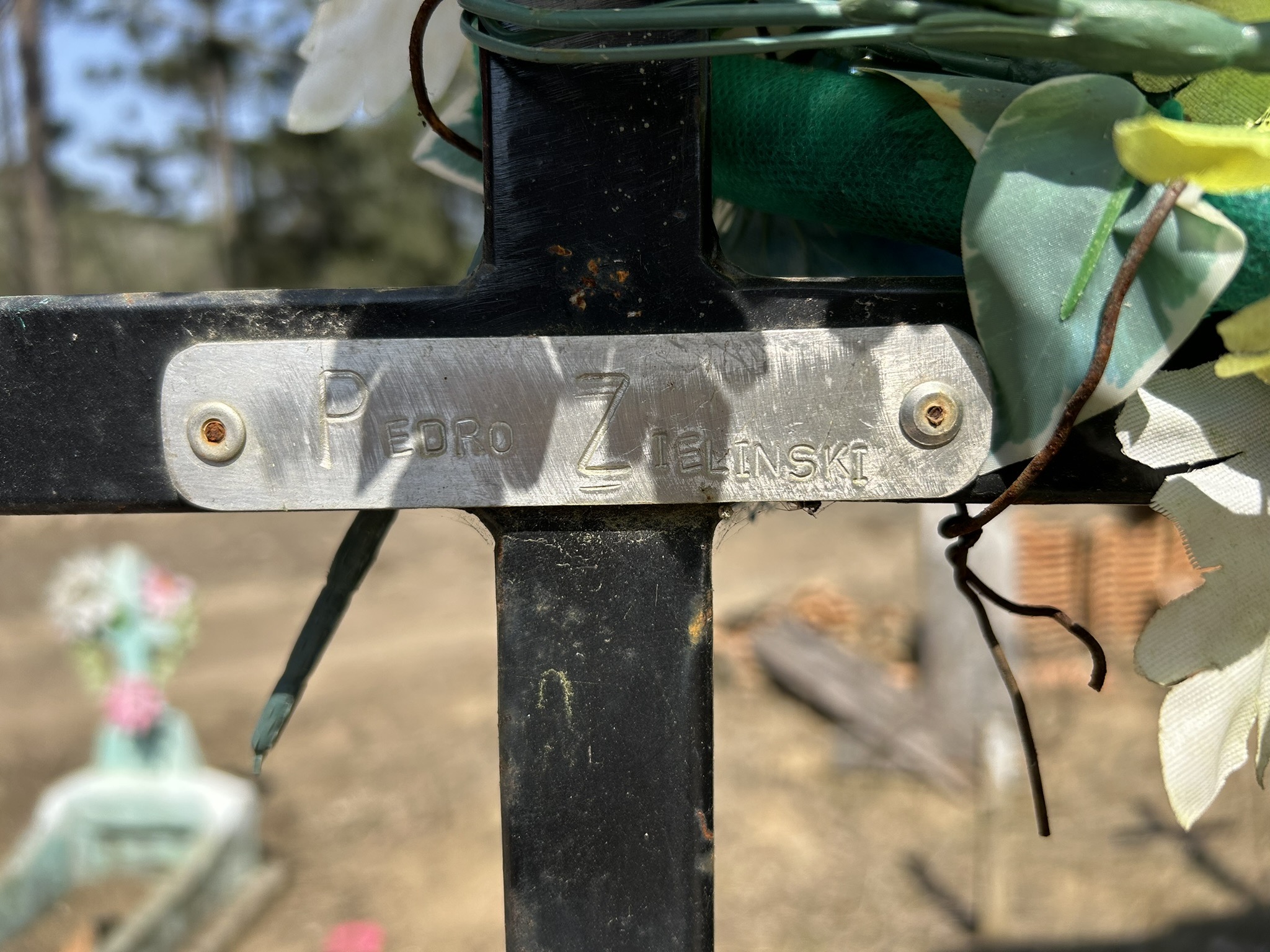 Gros plan d'une plaque métallique sur une tombe, sur laquelle est gravé le nom de "Pedro Zielinski". Autour de la plaque, des fleurs artificielles et du tissu vert.