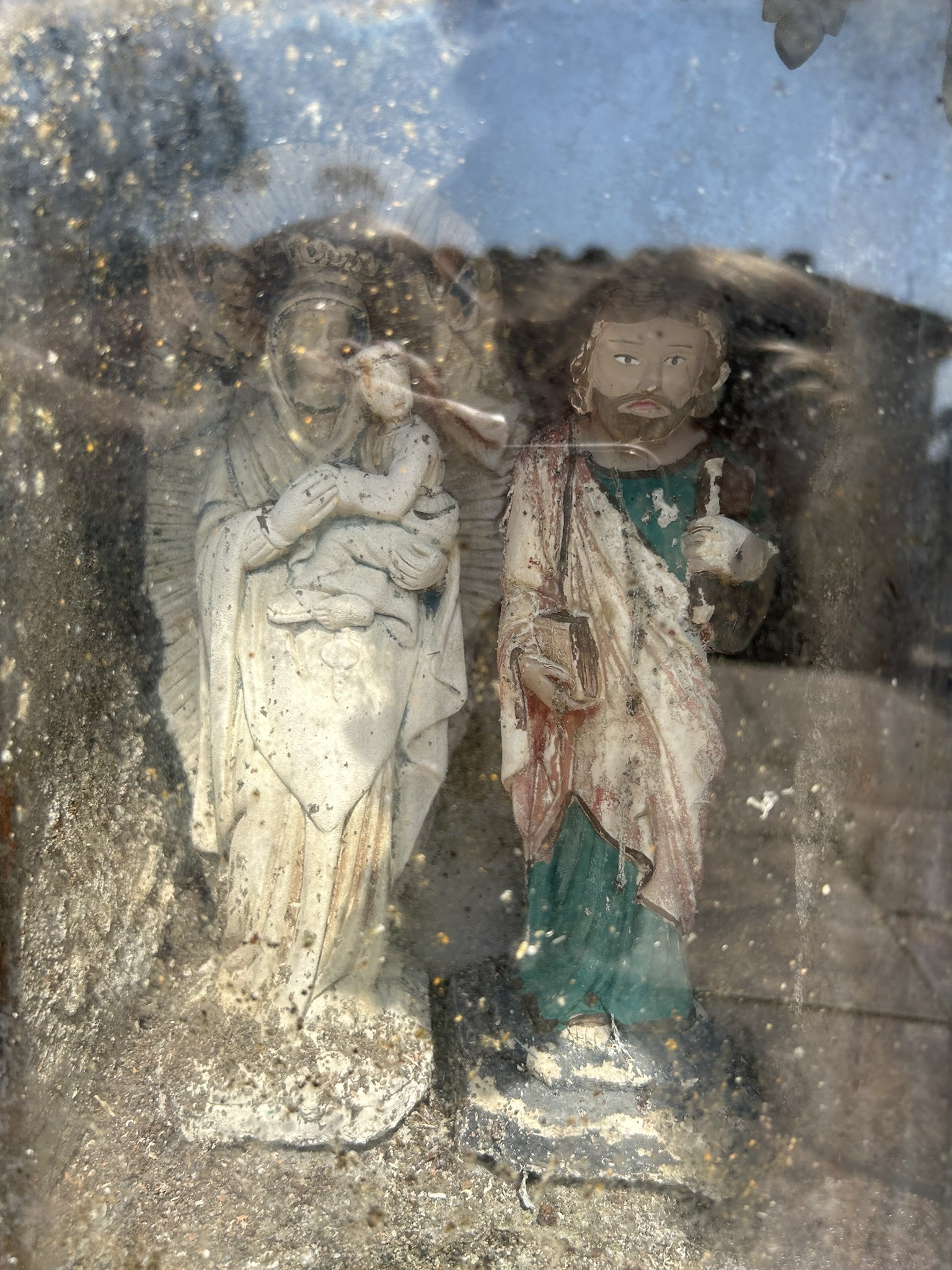 Figures religieuses détruites derrière le verre, l'une d'entre elles tenant un enfant, salissures et usures visibles.