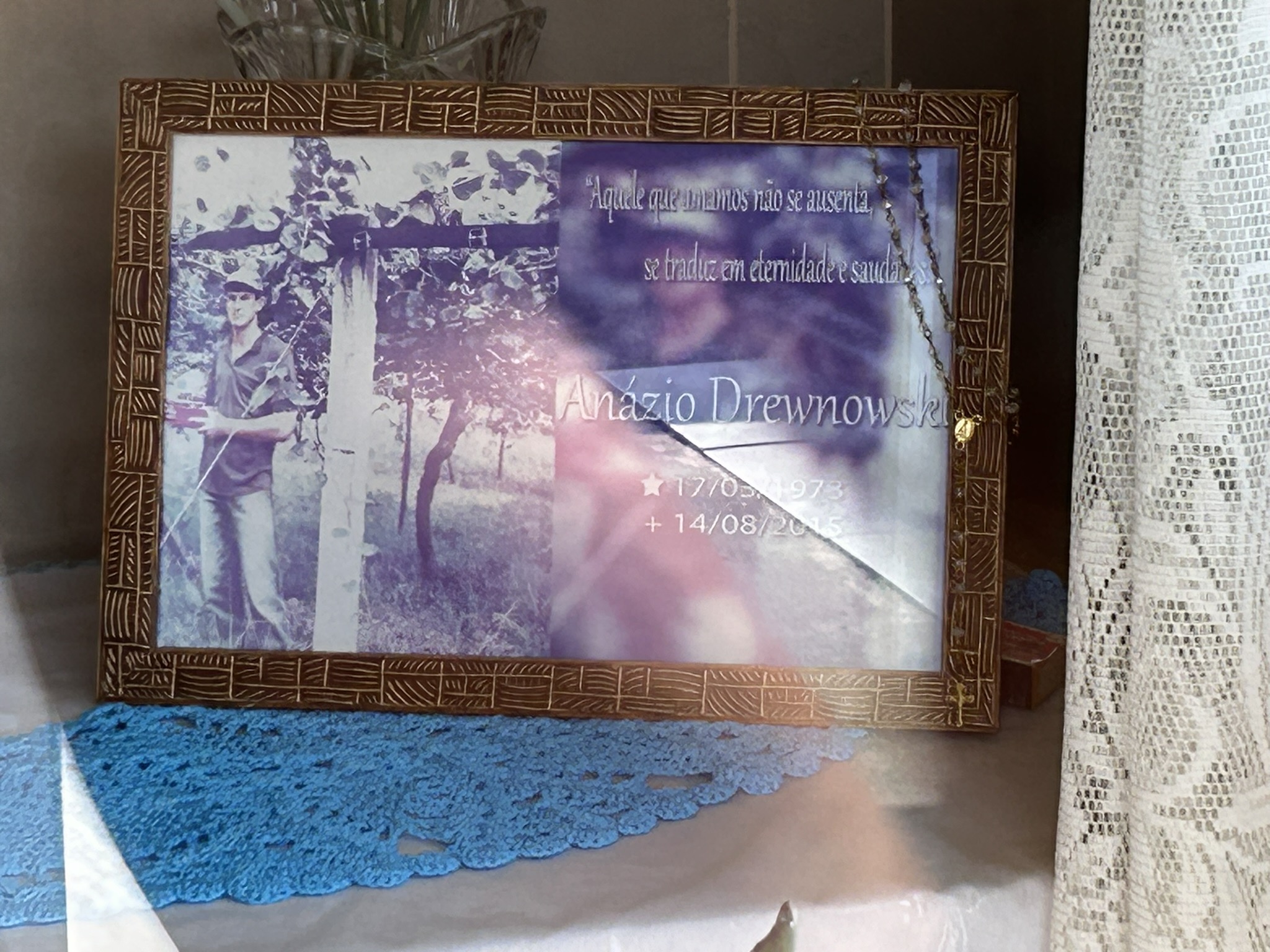 Framed photograph of a man standing in a vineyard, with text in Portuguese and the name Anazio Drewnowski, dates 17/03/1973 and 14/08/2015. The frame rests on a blue crochet napkin.