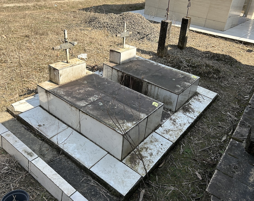 The grave of Regina and Zygmunt Michalski with two stone headstones and metal crosses on a tiled base, surrounded by dry grass.