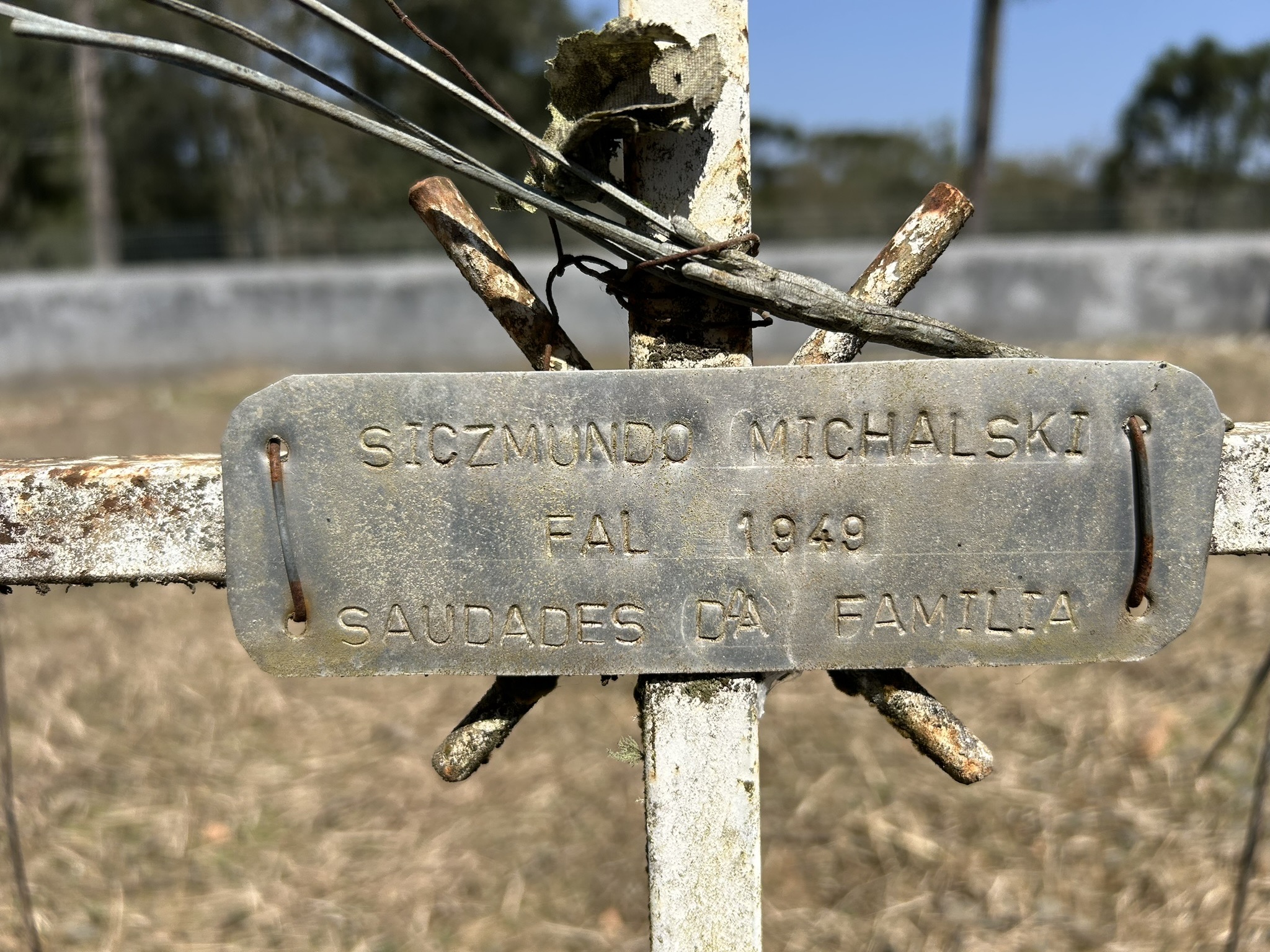 Metal plaque on grave with inscription: SICZMUNDO MICHALSKI, FALL 1949, SAUDADES DA FAMILIA. Plaque attached to a rusted metal cross.