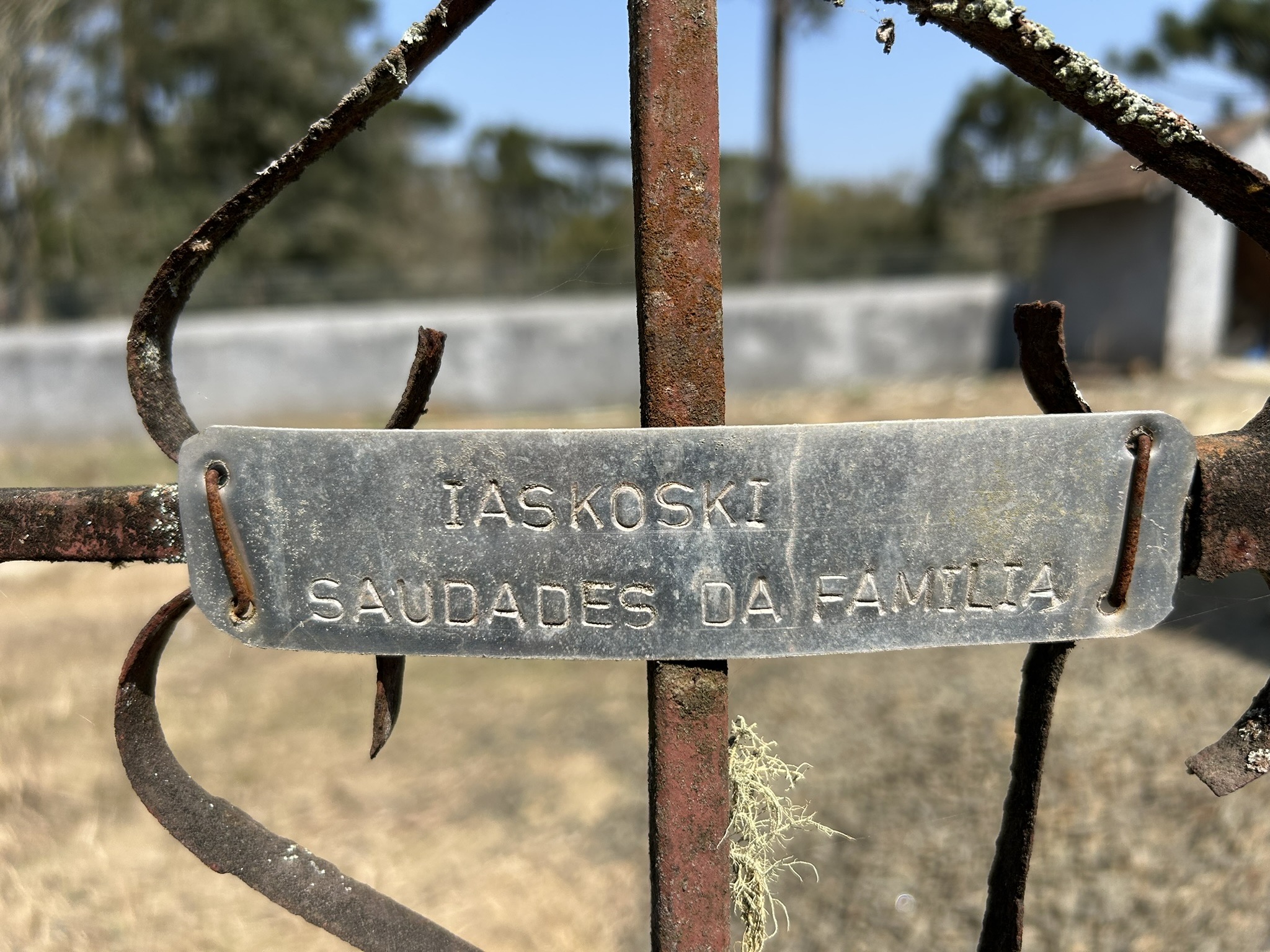 Металева табличка на іржавому паркані з написом "IASKOSKI SAUDADES DA FAMILIA" крупним планом.