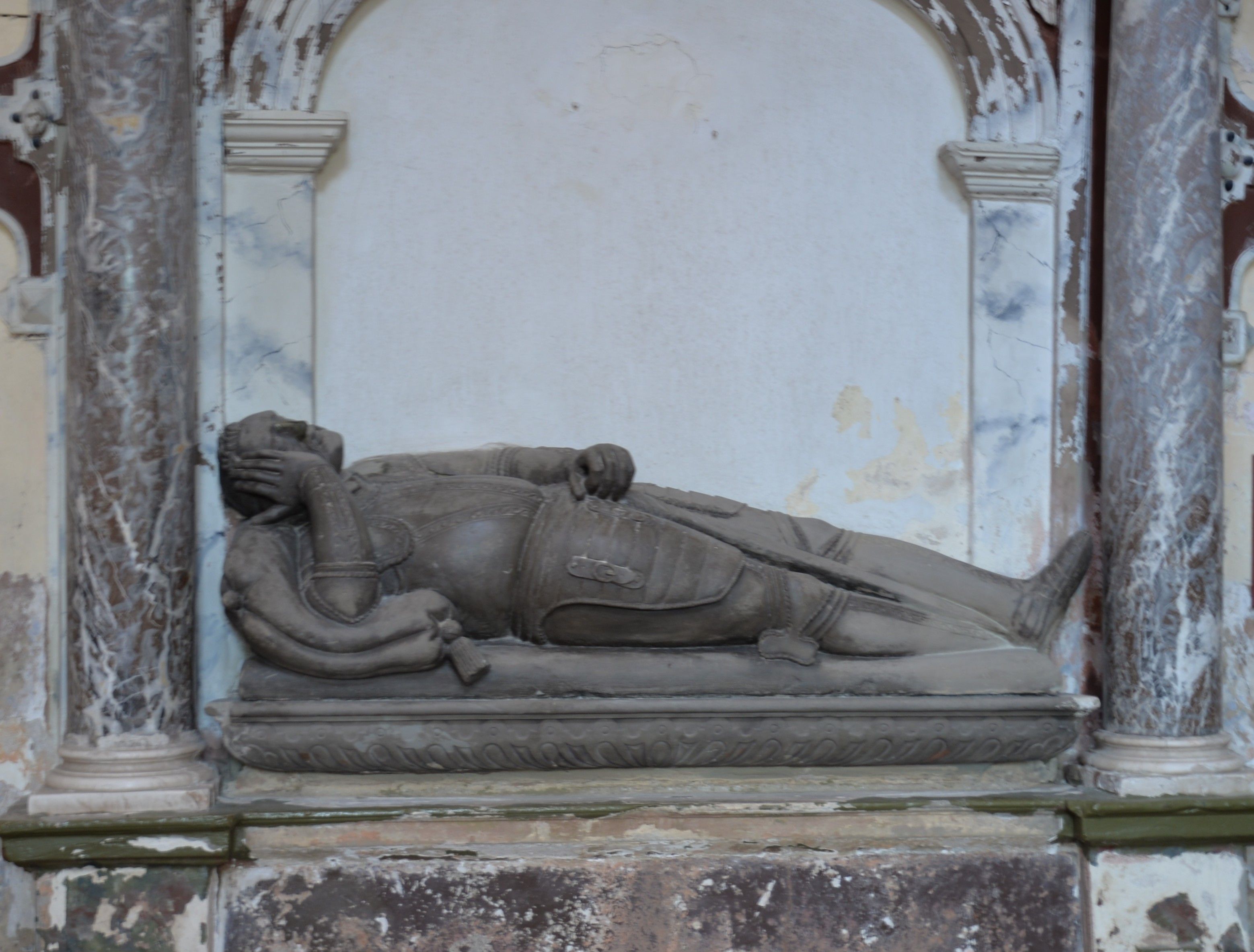 Stone effigy of a reclining knight in armor on a tomb, set against a decorative wall with marble columns. The knight rests his head on his hand.