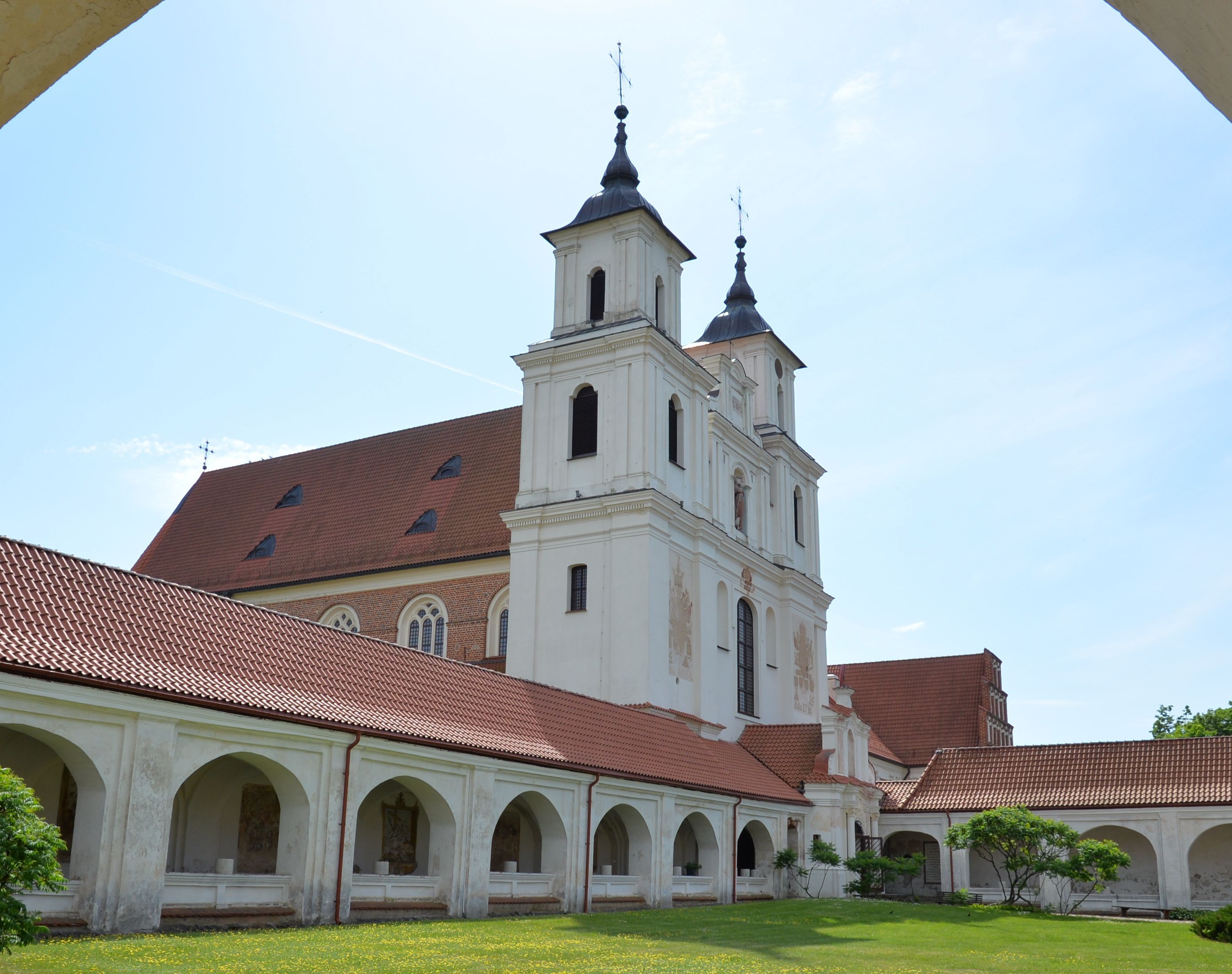 The Bernardine church and monastery in Cytowiany, featuring two towers and surrounded by arcaded cloisters with a red-tiled roof. The building is set against a clear blue sky.
