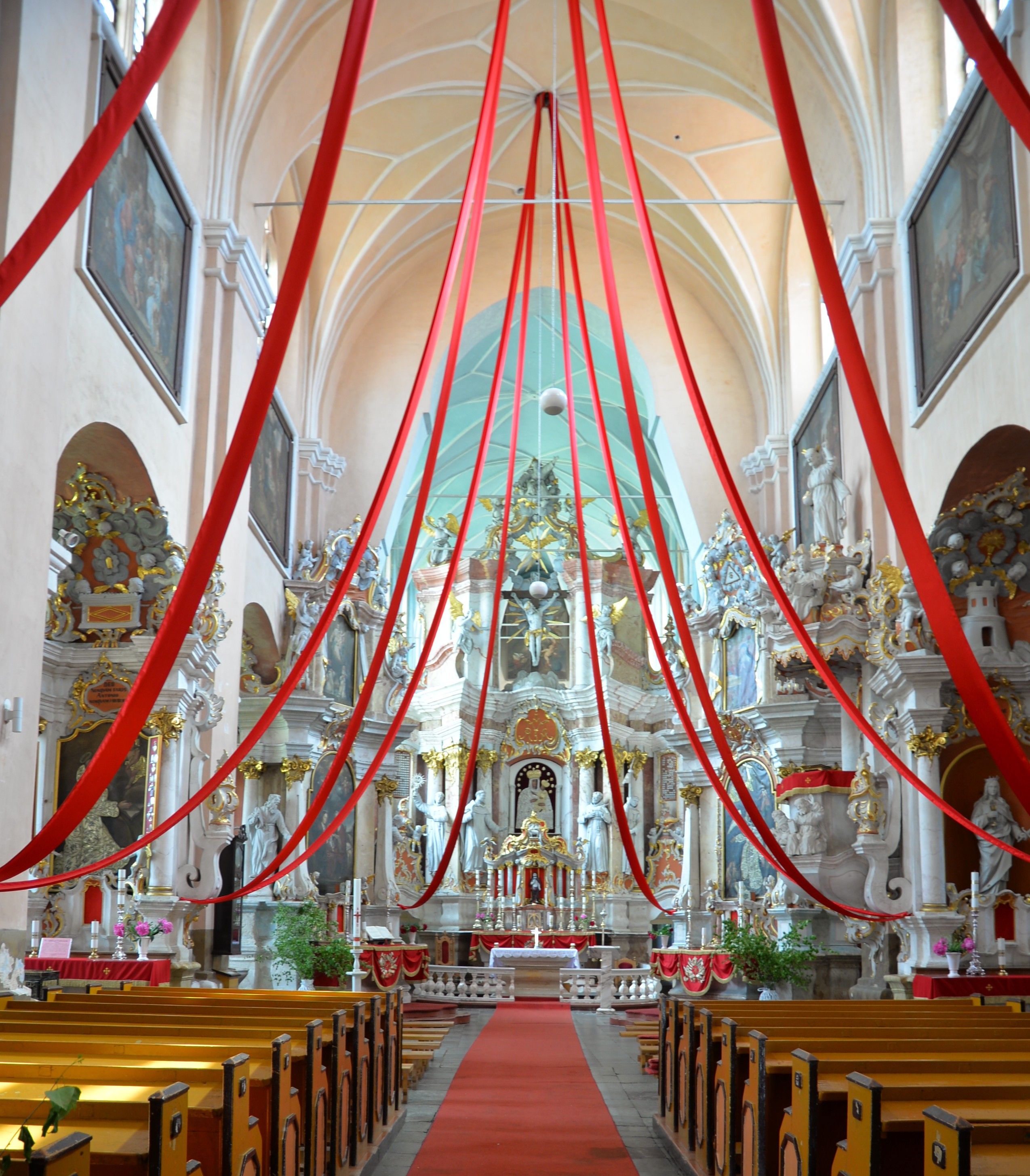 Interior of the Bernardine church in Cytowiany. The nave features red ribbons hanging from the ceiling, leading to an ornate altar with sculptures and paintings. Wooden pews line the aisle.