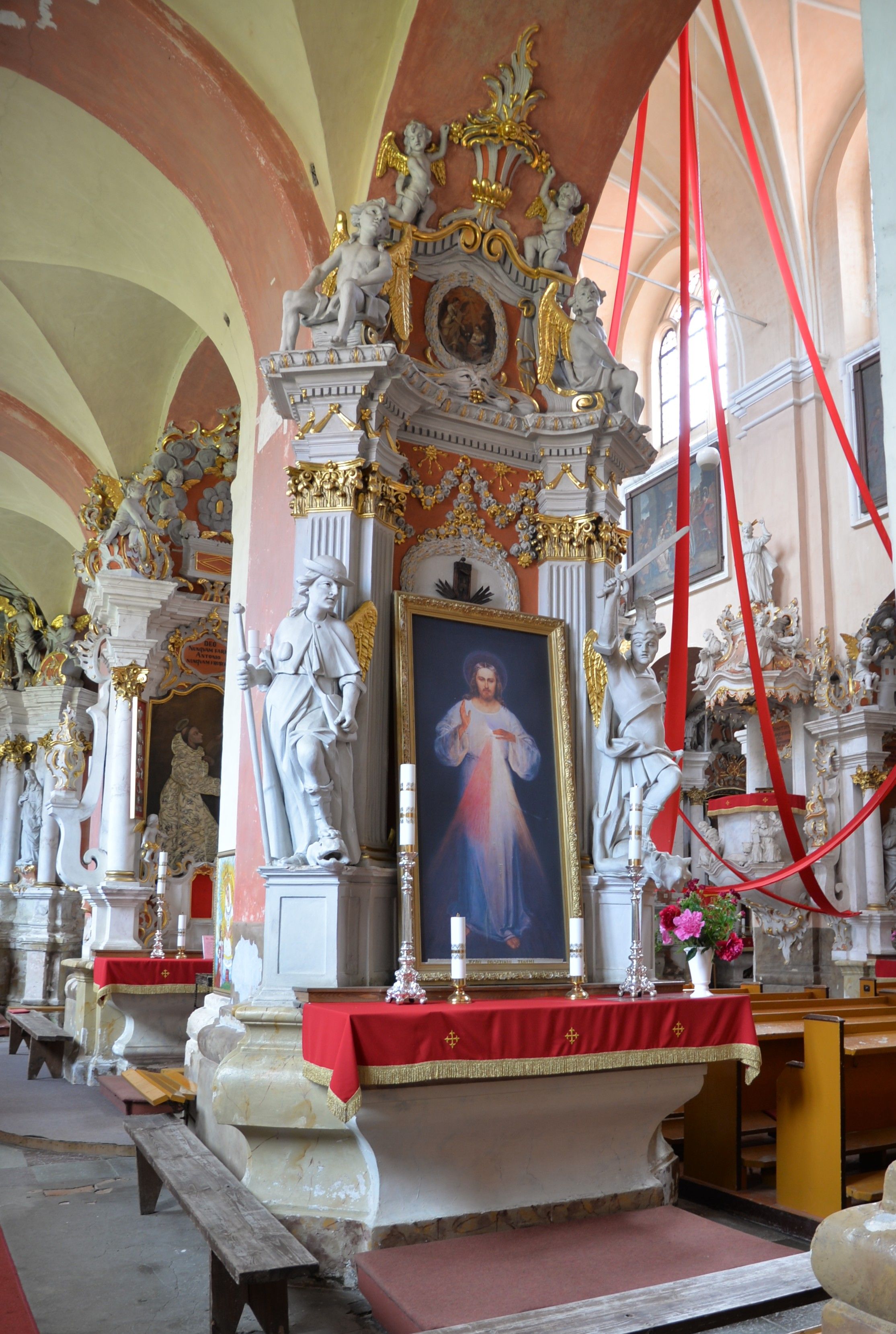 Interior of the Bernardine church in Cytowiany, featuring a richly decorated altar with a painting of Jesus. The altar is adorned with sculptures, gold accents, and red drapery.